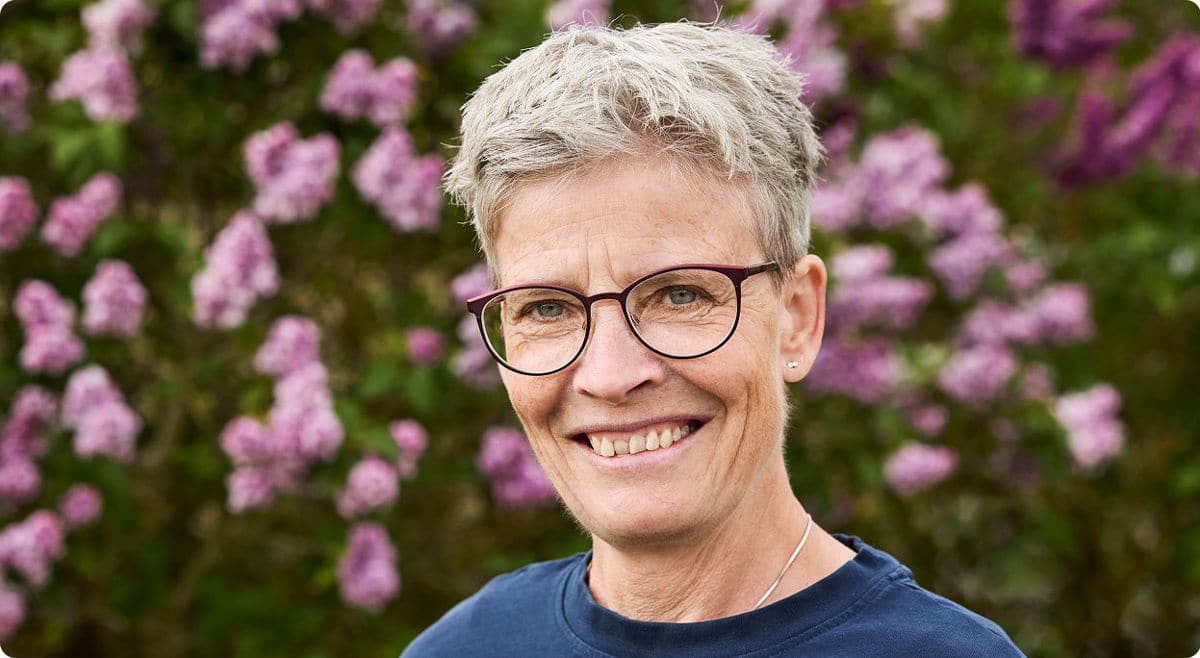 Frau mit kurzen Haaren und Brille lächelt und steht vor einem rosa blühendem Busch